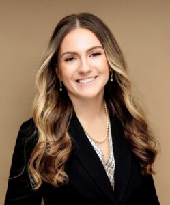 Kaitlin is posing in a professional headshot against a tan backdrop wearing a dark jacket and pearl necklace